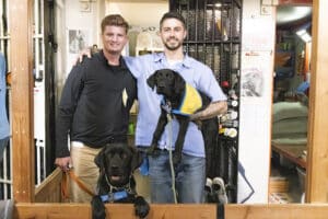 Two men pose together with two black Labrador dogs, one wearing a blue vest and the other in a yellow vest, in an indoor setting with various items in the background.