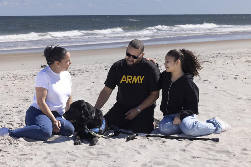 A family of three sitting on the beach with their black service dog wearing a blue vest, enjoying the ocean view in the background.