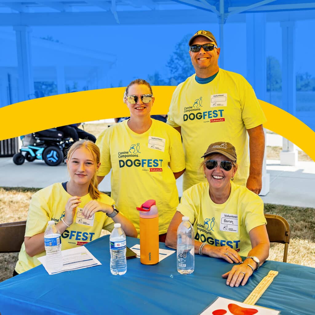 A group of four volunteers smiling at a table during a DogFest event, wearing matching yellow t-shirts with 'DogFest' written on them. They are seated behind a blue table, which has water bottles and papers on it, with a light background featuring a stroller in the distance.