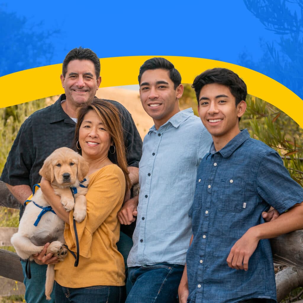 A cheerful family of four poses outdoors with a golden retriever puppy. The mother, wearing a yellow top and holding the puppy, is flanked by the father in a black shirt, and two sons in light blue and dark blue shirts, standing next to a wooden fence with a natural landscape in the background.