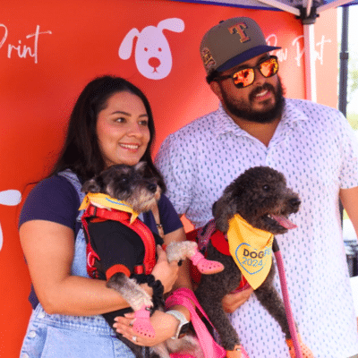 A woman and a man pose together under a tent, each holding a small dog dressed in colorful bandanas. The background features a graphic of a dog silhouette and the words 'Paw Print.'