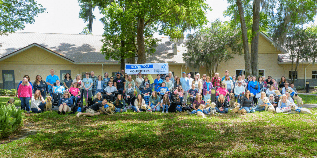 A large group of volunteers poses for a photo outdoors, holding a banner that reads 'Thank You Volunteers' for Canine Companions, with several service dogs present among them; a building is visible in the background, surrounded by trees and grass.