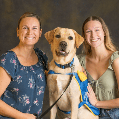 A smiling woman and a young woman pose together with a large, friendly-looking yellow Labrador Retriever. The dog is wearing a blue and yellow vest, and they are set against a dark, blurred background.