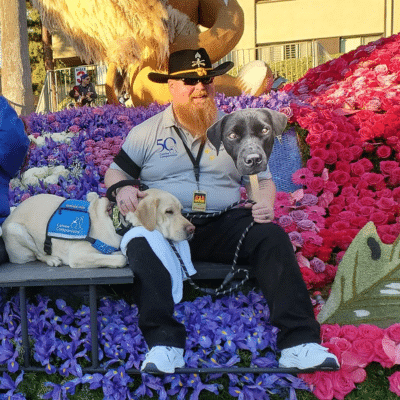 A man with a long red beard and a cowboy hat sits on a parade float surrounded by colorful flower decorations. He has a Labrador Retriever next to him and is holding a cardboard cutout of a black dog.