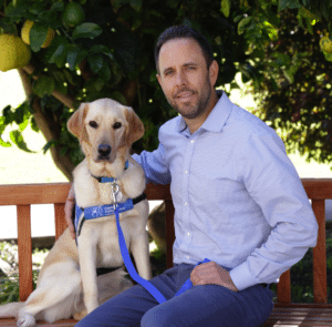 A man sitting on a wooden bench with a golden retriever beside him, both looking at the camera. The dog is wearing a blue harness with a leash attached, and there are green trees and lemons in the background.
